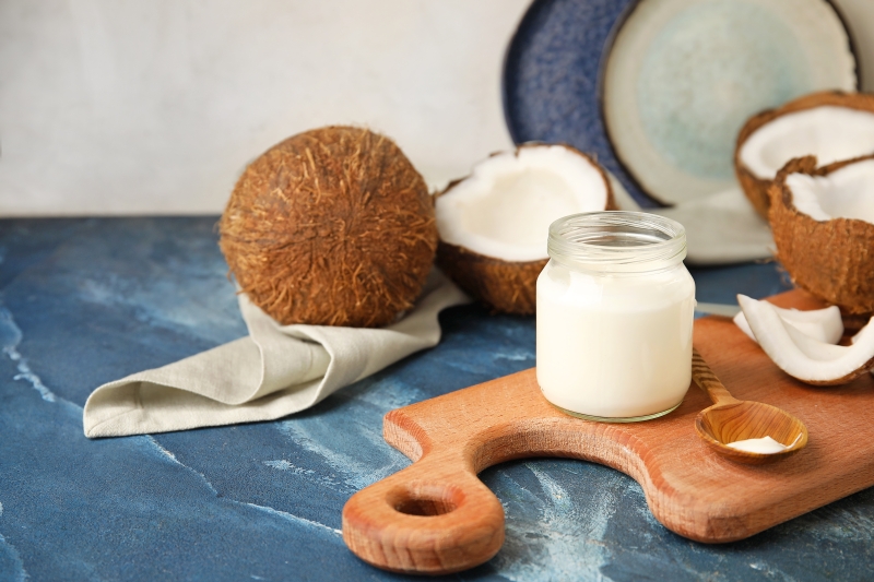 roślinny jogurt kokosowy w słoiczku na drewnianej desce, obok połówki orzecha kokosowego, ściereczka i łyżka